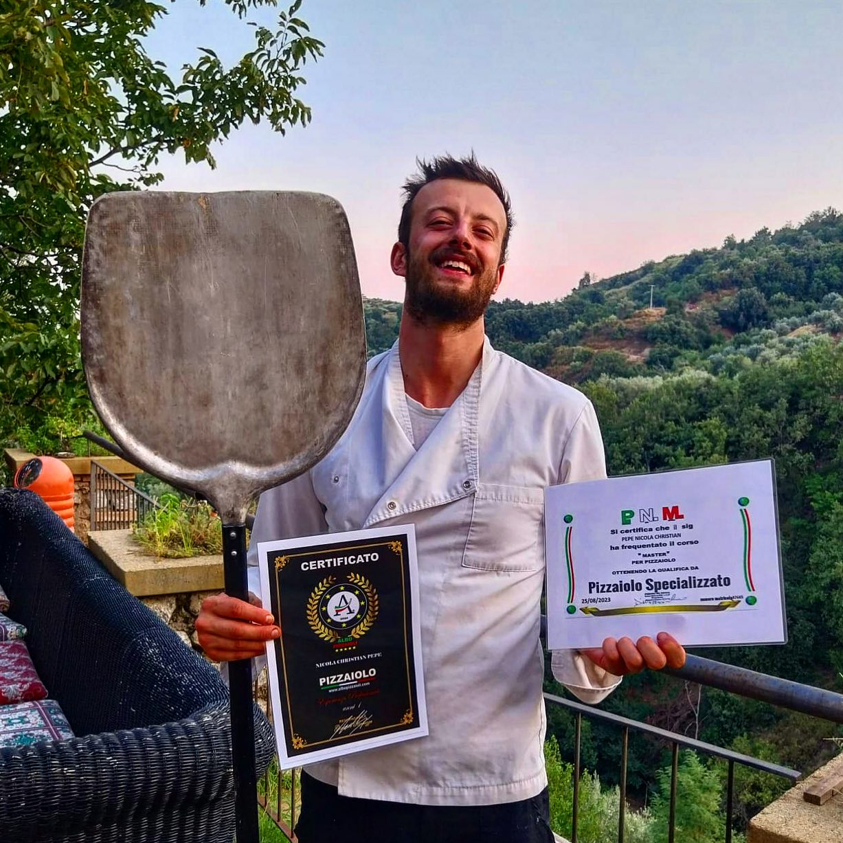 Nicola Pepe smiling outdoors while holding a pizza peel and certificates.