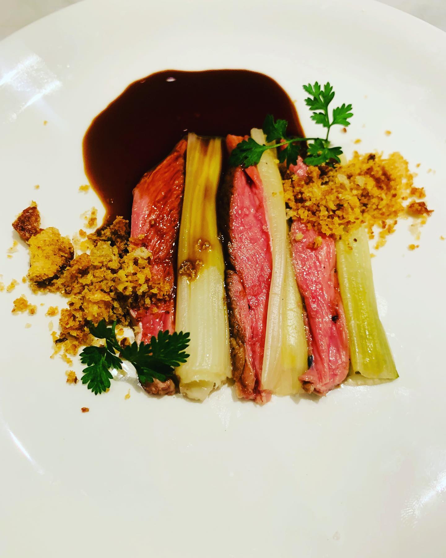 Refined plated meat dish with leeks, crumble, and sauce.